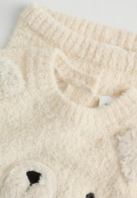 Kremowy, puszysty sweter o teksturowym wykończeniu. Posiada okrągłe, miękkie akcenty w formie aplikacji przypominających twarz oraz kontrastowe czarne detale.