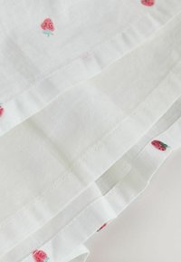 Tissu blanc avec de petits motifs de fraises rouges, présentant un design plissé et fini par un bord soigneusement cousu.