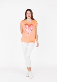 Camiseta de algodón naranja con un diseño de corazón colorido y el texto "PAZ". Mangas cortas, cuello redondo, combinada con jeans blancos y zapatillas.