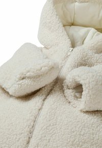 Veste en polaire couleur crème avec capuche, présentant une texture douce et duveteuse, une fermeture éclair frontale et des poignets surdimensionnés pour plus de chaleur.