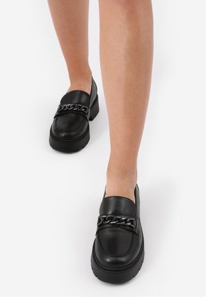 Sorte læderloafers med en kraftig sål og en dekorativ kæde hen over fronten. Har en glat tekstur og afrundet form.