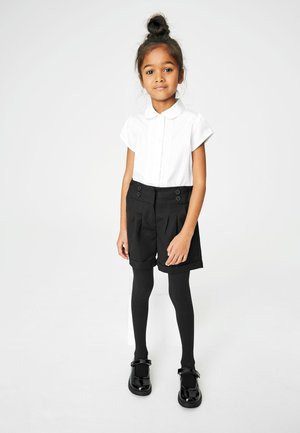 Camisa blanca de manga corta con cuello y botones decorativos, combinada con pantalones cortos negros de cintura alta con pliegues y dos botones frontales. Medias negras y zapatos brillantes negros completan el atuendo.