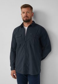 Camicia button-up blu scuro realizzata in tessuto morbido, con due tasche sul petto, colletto ampio e maniche lunghe. Indossata sopra una T-shirt bianca.
