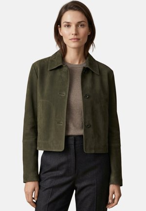 Woman wearing an olive green suede cropped jacket over a taupe sweater and dark gray tailored pants, standing against a plain background.