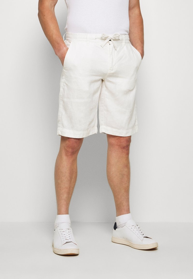 Homme portant un short blanc jusqu'aux genoux avec une taille à cordon, des chaussettes blanches et des baskets blanches, debout devant un fond uni.