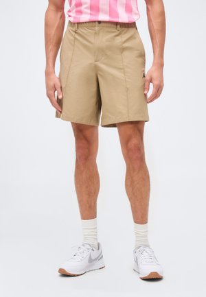 Mies, jolla on beige polvipituiset shortsit, valkoiset tennarit ja valkoiset sukat, seisoo yksivärisen taustan edessä.