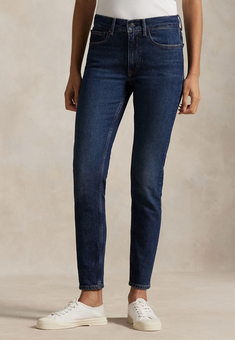 Persona che indossa jeans scuri attillati e sneaker bianche, in piedi contro uno sfondo beige neutro.