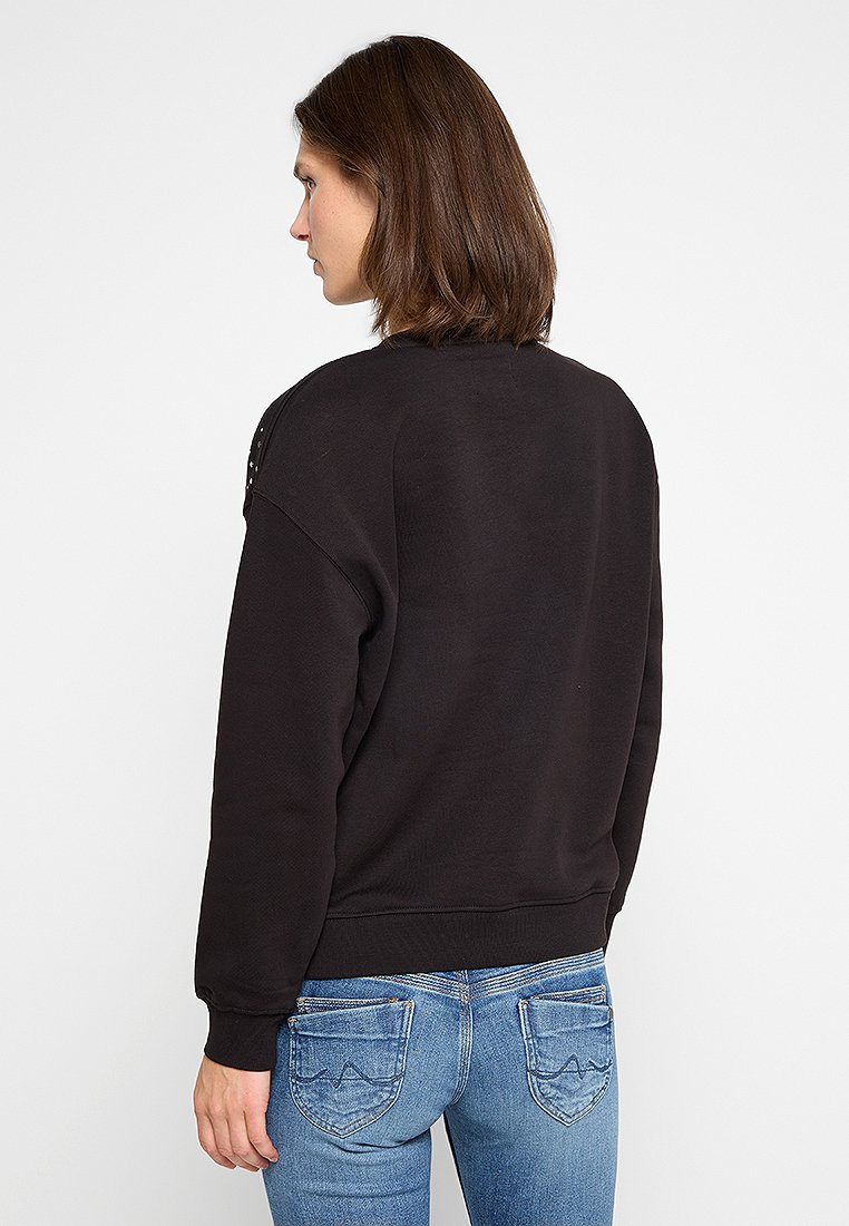 Sudadera negra con un corte holgado, tela texturizada y puños acanalados. Presenta detalles de tachuelas en los hombros. Se lleva con vaqueros azules.