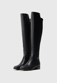 Un par de botas de cuero negras altas con tacones bajos y cremalleras laterales, de pie sobre un fondo liso.