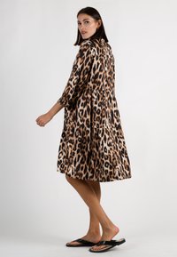 Kleid im Leopardenmuster aus leichtem Stoff, mit mehrlagigem Design, Dreiviertelärmeln und tailliertem Schnitt. Getragen mit schwarzen Flip-Flops.