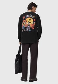 Camisa negra de manga larga con un gráfico colorido en la espalda que muestra una cara sonriente y patrones florales, combinada con pantalones negros y una bolsa.