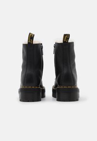 Bottes noires en cuir à la cheville avec une fermeture éclair à l'arrière, une doublure blanche douce et des coutures jaunes sur une semelle noire épaisse et texturée.