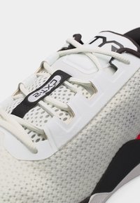 Κοντινό πλάνο λευκού αθλητικού παπουτσιού από δίχτυ με μαύρες, λευκές και κόκκινες λεπτομέρειες, που διαθέτει ελαστικά κορδόνια και μαύρη θηλιά έλξης με την επιγραφή «EXT-2».