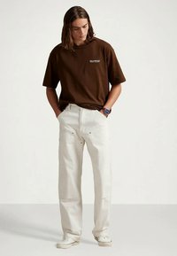 Młody mężczyzna z długimi włosami, ubrany w brązowy t-shirt Polo Sport, białe spodnie, białe sneakersy i niebieski zegarek na ręku, stoi z rękami w kieszeniach.