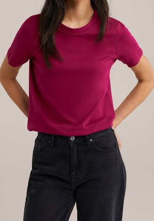 Enganliegendes T-Shirt mit kurzen Ärmeln aus leuchtend magentafarbenem Baumwollstoff, runder Ausschnitt, glatte Textur, kombiniert mit hochgeschnittenen schwarzen Jeans.