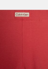 Crvene rebraste tajice s bijelom etiketom na kojoj piše "Calvin Klein" smeštenom blizu struka. Minimalistički dizajn bez uzoraka.