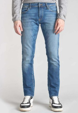 Jeans en denim bleu clair avec une coupe slim, lavage moyen et légères décolorations. Style classique à cinq poches, associés à des baskets noires et blanches.