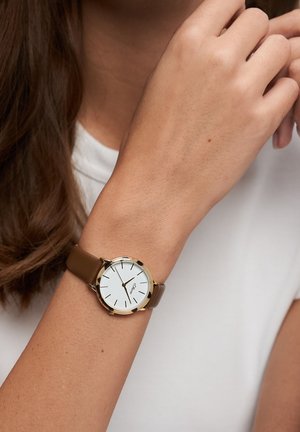 Montre en cuir marron avec un boîtier en métal doré, cadran blanc, indices horaires noirs et design minimaliste, portée au poignet.