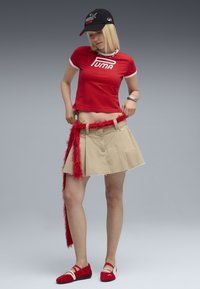 Röd Puma kortärmad t-shirt, beige kjol med rå fåll, lurvig röd bälte och röda platta skor med korsade remmar. Svart Puma keps.