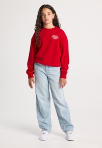 Ragazza con lunghi capelli ricci che indossa una felpa rossa con la scritta "La Città dei Sogni New York", jeans azzurri e sneakers bianche in piedi.