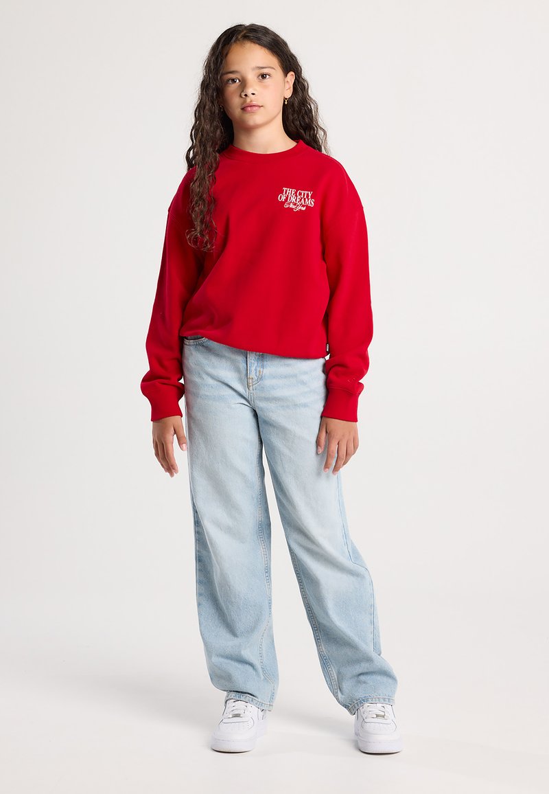 Ragazza con lunghi capelli ricci che indossa una felpa rossa con la scritta "La Città dei Sogni New York", jeans azzurri e sneakers bianche in piedi.