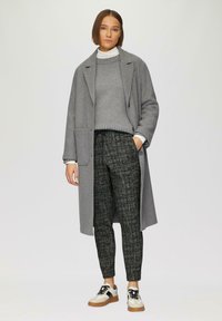 Grijze wollen jas over een grijze trui, gestructureerde zwart-grijze patroonbroek, en witte sneakers met zwarte accenten en bruine zolen.