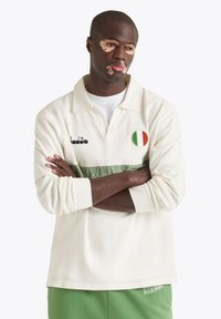 Polo a maniche lunghe di colore crema con una striscia orizzontale verde, dotato di logo e patch della bandiera italiana. Tessuto morbido e vestibilità rilassata.