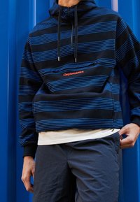 Felpa con cappuccio a strisce nere e blu con tasca frontale. Presenta un cappuccio con coulisse e il logo arancione "Clepomanicx". Indossata sopra pantaloncini grigi.