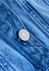Zbliżenie na niebieski materiał dżinsowy z srebrnym guzikiem oznaczonym "NEXT ESSENTIAL JEANSWEAR" oraz widocznymi detalami szwów.