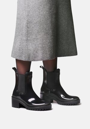 Bottines noires brillantes à talons épais portées avec des collants noirs et une jupe grise jusqu'aux genoux sur un fond uni.