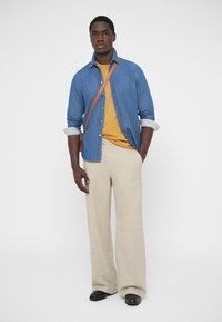 Džinsinis marškinėlis dėvimas virš geltonos palaidinės, kartu su smėlio spalvos platėjanciais kelnais ir juodais batais; komplektuojama su rudos spalvos rankinės dirželiu per petį.