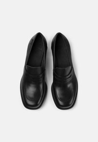 Mocassins en cuir noir avec une texture lisse, présentant un bout arrondi et des coutures décoratives à l'avant. Semelle plate, sans matériel visible.