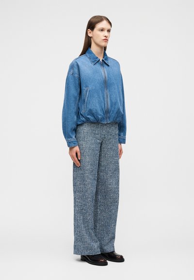 Femme debout portant une veste en denim bleue zippée, un pantalon bleu texturé à jambes larges et des chaussures marron foncé sur un fond clair uni.