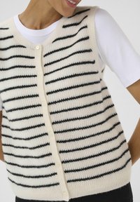 Persona sorridente che indossa un gilet a righe di colore crema sopra una camicia bianca, mostrando i bottoni e la texture su uno sfondo neutro.