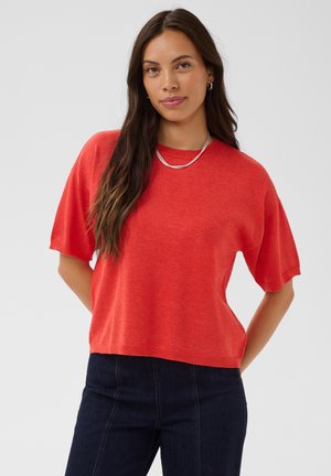 Femme aux longs cheveux bruns portant un pull rouge à manches courtes et un jean foncé, debout les mains dans le dos devant un fond uni.