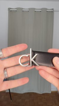 Črni usnjeni pas s srebrnim zaponkom v obliki črk "CK", ki ga držita dve roki. Mehka tekstura, eleganten dizajn in minimalni detajli.