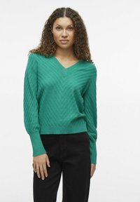 Grüner Strickpullover mit V-Ausschnitt, der ein Zickzackmuster aufweist und lange Ärmel hat. Kombiniert mit schwarzen Hosen für einen Kontrast.