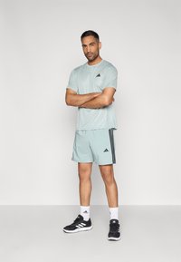 Svetlo turkizna športna majica in kratke hlače z črnimi logotipi Adidas in tremi črnimi črtami. Črni športni copati, bele nogavice. Gladek tekstura.