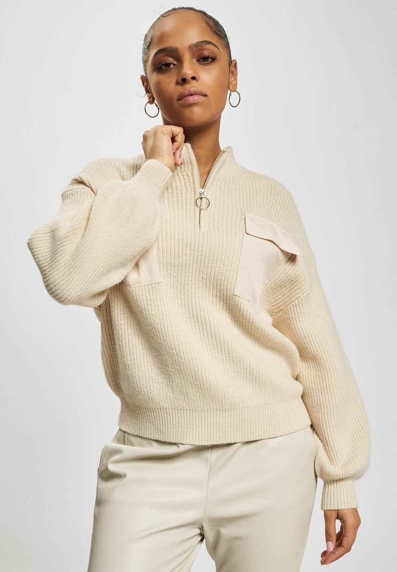 Beige strikkegenser med glidelås i kragen, med to lyse lommer foran. Designet har ribbestruktur og vide ermer.
