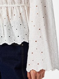 Haut blanc à manches longues avec broderie florale en œillets et bords festonnés, porté sur un jean denim bleu foncé.