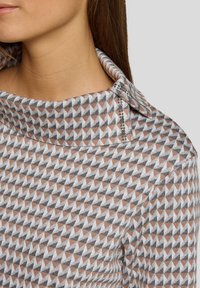 Frau in einem hochgeschlossenen Oberteil mit einem geometrischen Dreiecksmuster in Grau, Weiß und Beige, das nahe dem Kragen mit kleinen dekorativen Pailletten verziert ist.