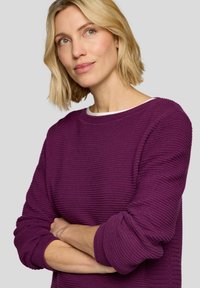 Lila strukturierter Pullover mit rundem Ausschnitt und langen Ärmeln, der ein geripptes Muster und umgeschlagene Bündchen aufweist. Der Hintergrund ist neutral grau.