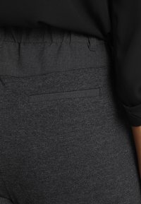 Pantalon extensible gris avec taille élastiquée et une poche arrière discrète. La texture du tissu semble douce et légèrement chinée.