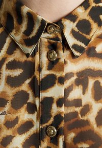 Gros plan d'un col de chemise à imprimé léopard avec trois boutons décoratifs en or présentant un motif de tête de lion.