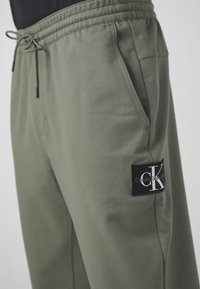 Olivgröna träningsbyxor i bomullsblandning med elastisk midja, dragsko, sidofickor och en svart logotypapplikation på nedre ben.