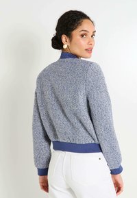 Une femme aux cheveux bruns relevés en chignon porte une veste bleue texturée avec des poignets et un col côtelés, assortie à un pantalon blanc, se retournant par-dessus son épaule.