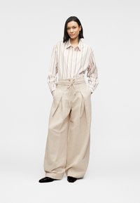 Μπεζ παντελόνι φαρδύ στο πόδι με πιέτες και ζώνη με κορδόνι στη μέση, συνδυασμένο με λευκό πουκάμισο με παστέλ κάθετες ρίγες. Μαύρα παπούτσια.