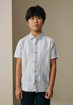 Chlapec s rovnými čiernymi vlasmi, oblečený v bielej krátkorukávovej košeli s modrými vertikálnymi pruhmi a tmavými nohavicami, stojaci na neutrálom pozadí.