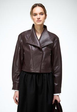 Jonge vrouw met een donkergroene leren cropped jas en een zwarte rok, die een pluizige donkergroene handtas vasthoudt tegen een witte achtergrond.