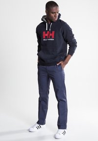 Sudadera azul marino con un logo rojo y blanco, forro de capucha gris, combinada con pantalones azul oscuro y zapatillas blancas con rayas negras.